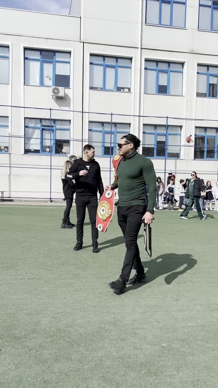 Antrenorul traversează terenul de sport al unei școli cu centura de campion pe umăr, spre elevii aliniați pe margine.