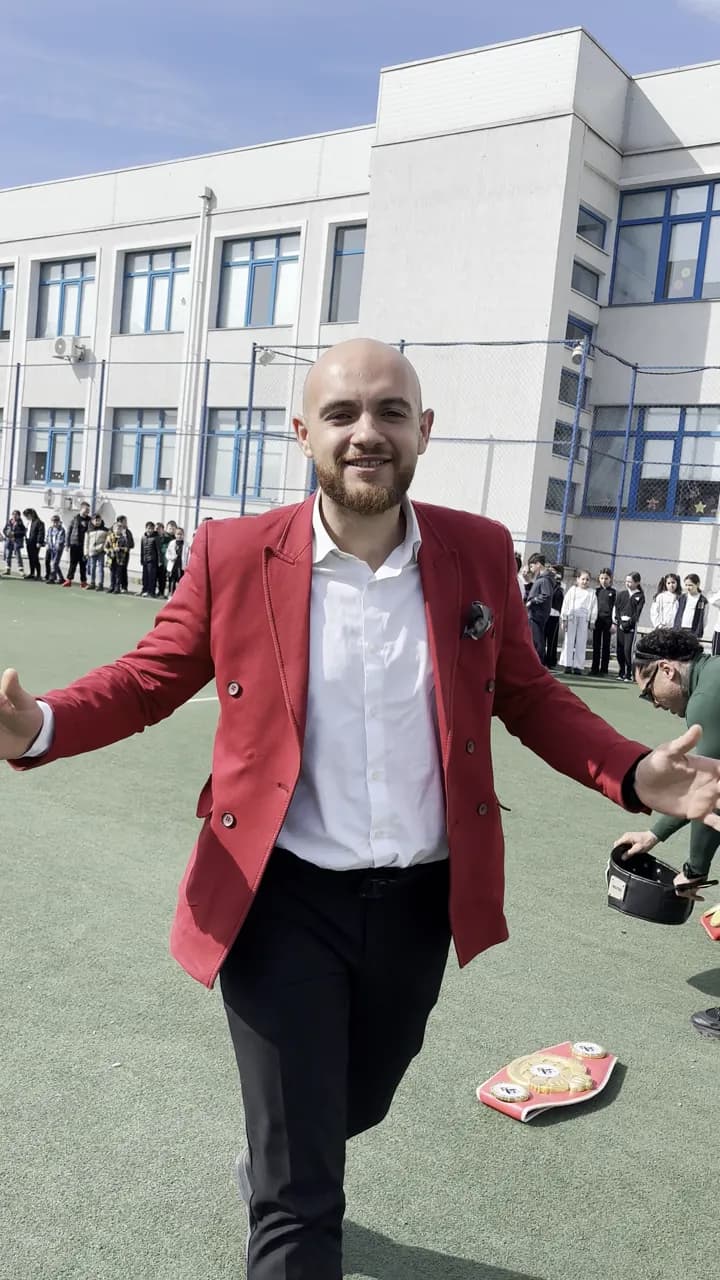 Un bărbat în sacou roșu vorbește către cameră pe terenul școlii, cu centura de campion așezată pe iarbă lângă el.