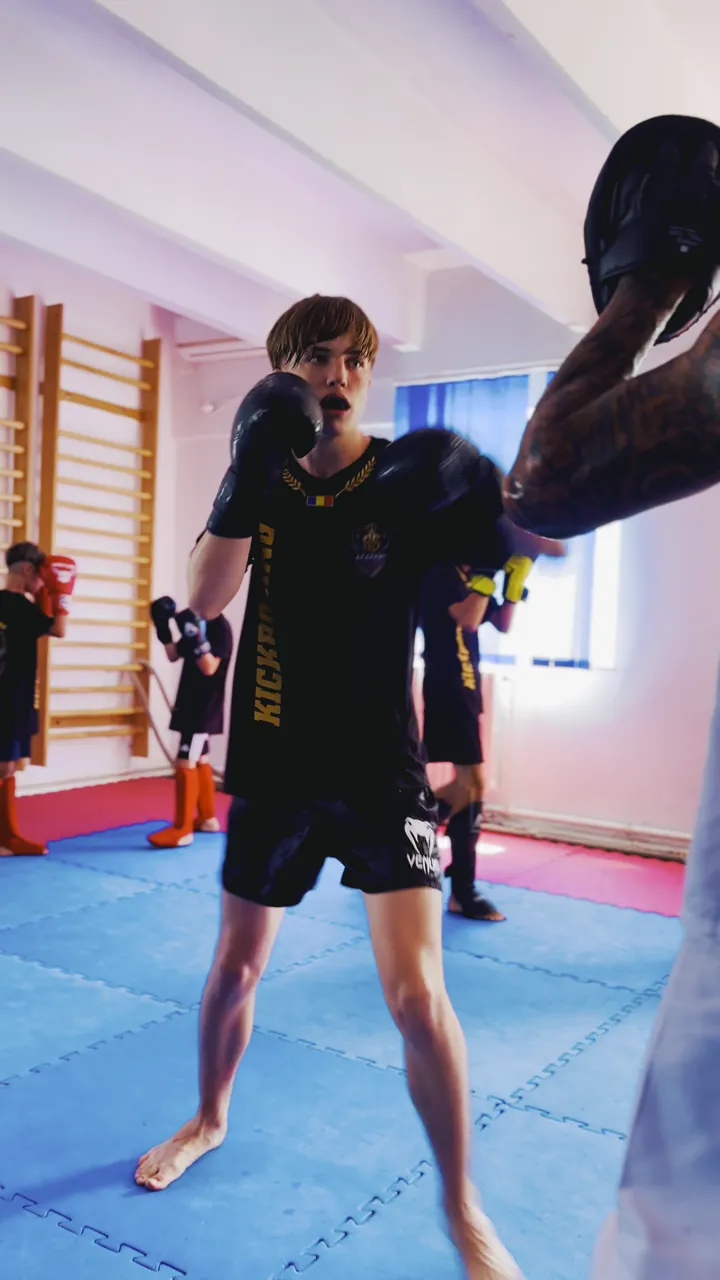 Adolescent cu garda sus pe tatami, cu antrenorul în fața lui; în fundal lucrează restul grupei, lângă spaliere.