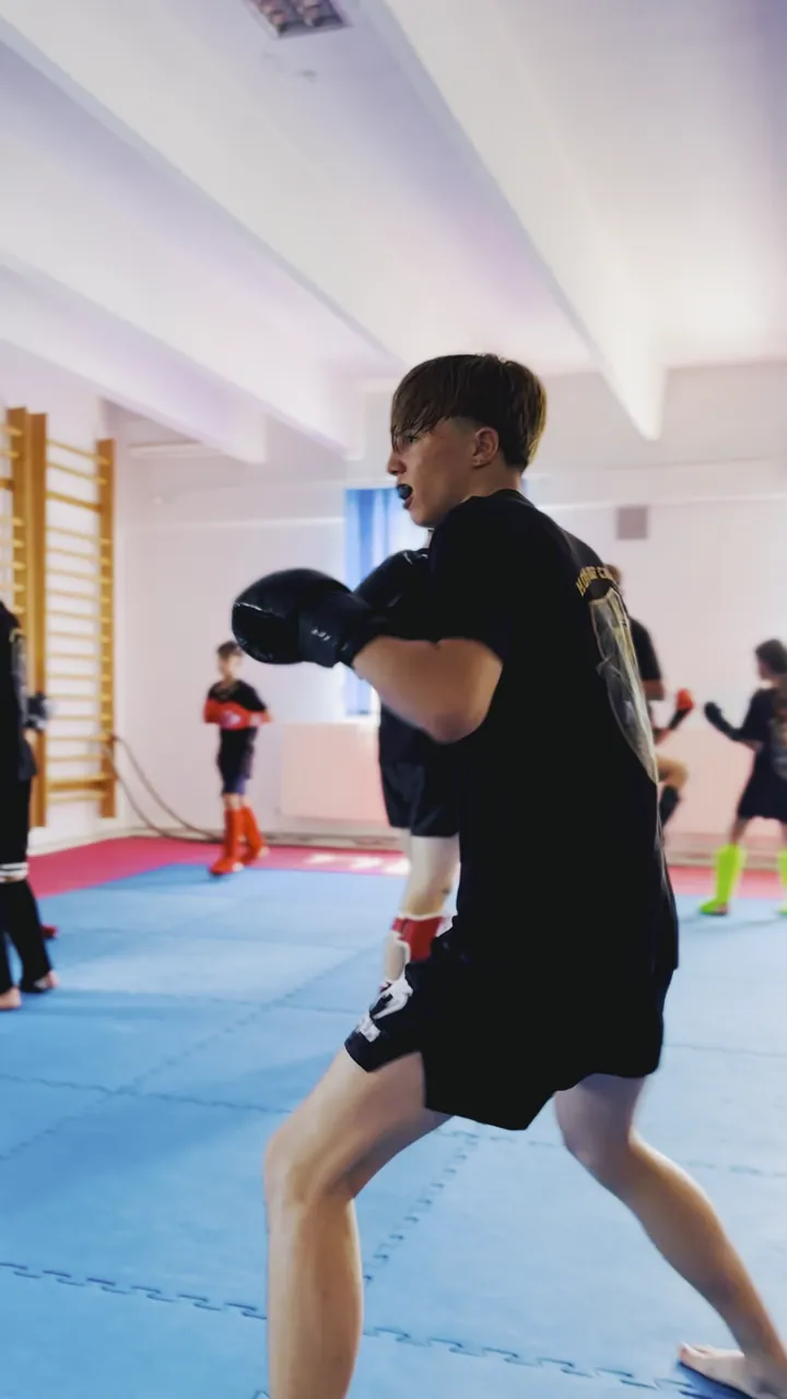 Lucru în perechi pe tatami, cu mănuși și protecții, într-o sală cu spaliere și saci de box.